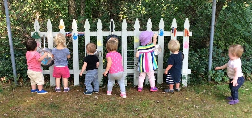 Toddlers making music with pots and pans on a fence